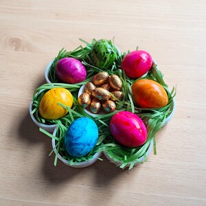 May include: A flower-shaped serving dish filled with colorful Easter eggs and chocolate candies. The eggs are dyed in shades of pink, yellow, blue, and orange, nestled in green paper grass. A small bowl in the center holds gold-wrapped chocolate eggs.