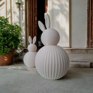 May include: Two white decorative bunny sculptures. The larger bunny has a round head and a ribbed, spherical body. The smaller bunny has a similar design. Both have long, upright ears. They are placed on a concrete surface near a potted plant.