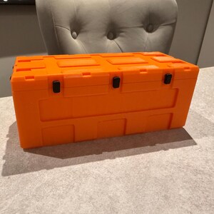 May include: Bright orange rectangular storage container with a textured surface. The container has a hinged lid and black latches. The container is sitting on a light gray surface.