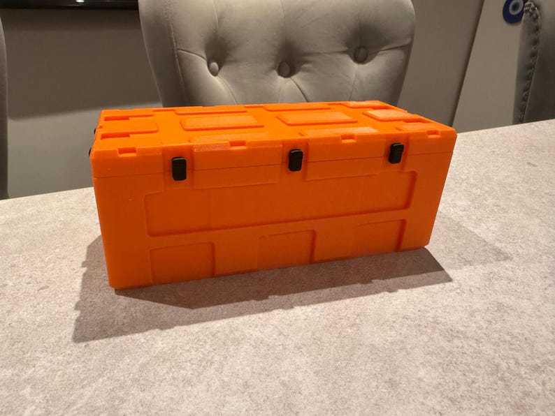 May include: Bright orange rectangular storage container with a textured surface. The container has a hinged lid and black latches. The container is sitting on a light gray surface.