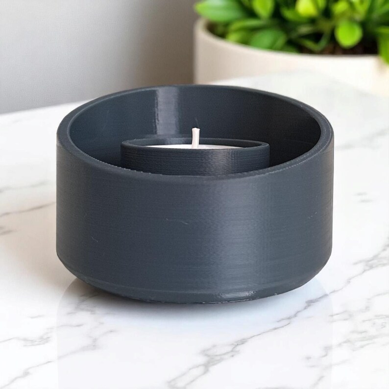May include: A dark gray candle holder with a circular design. The holder has a recessed center that holds a white tea light candle. The candle has a white wick. The holder is sitting on a white marble surface.