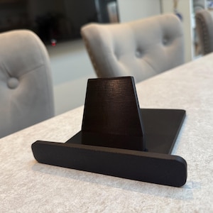 May include: A black wooden phone stand with a rectangular base and a vertical support. The stand is designed to hold a phone or tablet. The stand is on a light-coloured surface, with two grey upholstered chairs in the background.