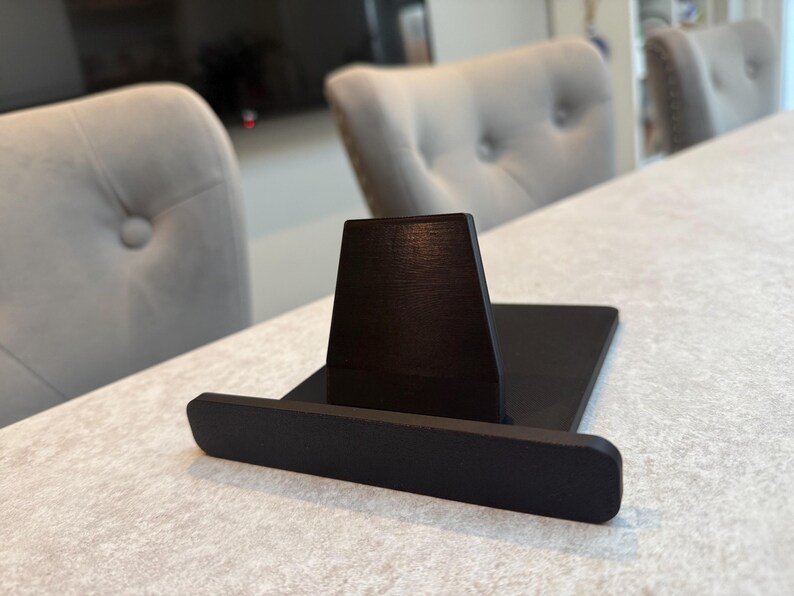 May include: A black wooden phone stand with a rectangular base and a vertical support. The stand is designed to hold a phone or tablet. The stand is on a light-coloured surface, with two grey upholstered chairs in the background.