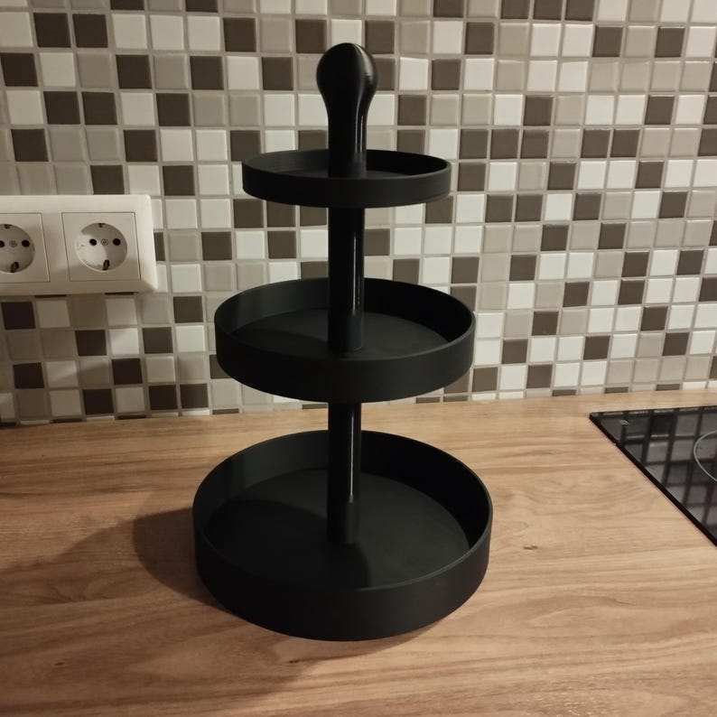 May include: A black, three-tiered serving tray with a central pole and a handle on top. The tray has three circular platforms of varying sizes, perfect for displaying food or decorative items. The tray is made of metal and sits on a wooden surface.