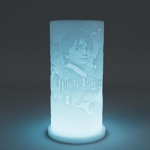 May include: A cylindrical, illuminated lamp featuring a portrait of Harry Potter with the text "Harry Potter". The lamp has a light blue glow and is set against a dark background. The design is etched into the lamp's surface.