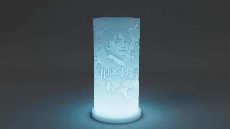 May include: A cylindrical, illuminated lamp featuring a portrait of Harry Potter with the text "Harry Potter". The lamp has a light blue glow and is set against a dark background. The design is etched into the lamp's surface.