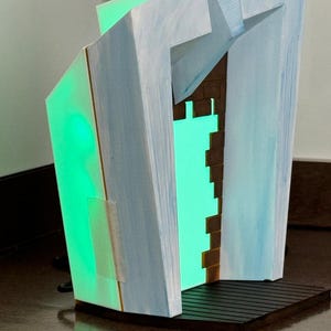 May include: Abstract geometric sculpture with a green internal light. The sculpture has a light blue and white exterior with a brown brick-like interior. The base is dark brown with a slatted design. The sculpture is illuminated with a bright green light.