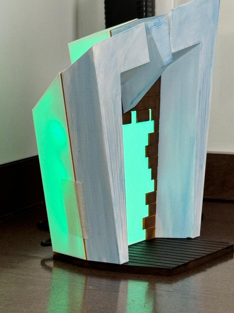 May include: Abstract geometric sculpture with a green internal light. The sculpture has a light blue and white exterior with a brown brick-like interior. The base is dark brown with a slatted design. The sculpture is illuminated with a bright green light.