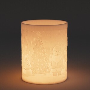 May include: A cylindrical, off-white Christmas candle holder with a festive design. The design includes a Christmas tree, Santa Claus, and a snowman, all set against a snowy landscape. The candle holder is illuminated from within, creating a warm glow.