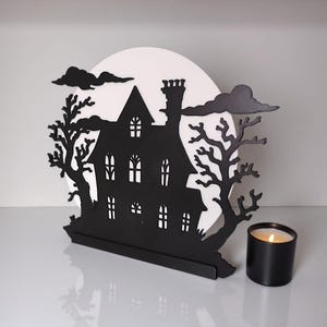 May include: A black silhouette of a haunted house with a white moon backdrop, trees, and clouds. A black candle with a lit wick sits to the right. The house has multiple windows and a chimney, creating a spooky Halloween decoration.