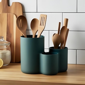 May include: A teal utensil holder with three sections, filled with wooden spoons and spatulas. The holder is on a wooden surface, with a white-tiled wall behind it. A wooden chopping board and a jar of flour are in the background.