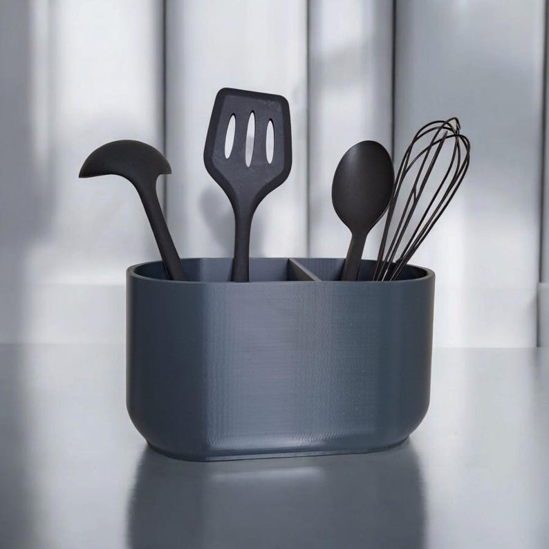 Puede incluir: Un soporte para utensilios de cocina gris oscuro con una variedad de utensilios de cocina negros. El soporte contiene un cuchar&oacute;n, una esp&aacute;tula ranurada, una cuchara y un batidor. El soporte est&aacute; dividido en tres secciones.