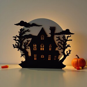 Pu&ograve; includere: Silhouette nera di una casa infestata con finestre illuminate, alberi e nuvole, su una luna bianca. Una zucca arancione e caramelle sono sul tavolo, creando un tema di Halloween.