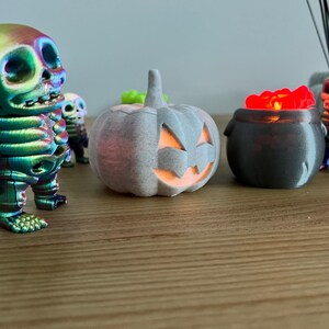May include: A Halloween-themed display featuring a rainbow-colored skeleton figurine, a gray pumpkin with a carved face, and a cauldron filled with glowing red objects. Additional skeleton figurines are in the background. The items are on a wooden surface.