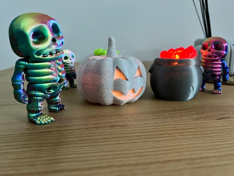 May include: A Halloween-themed display featuring a rainbow-colored skeleton figurine, a gray pumpkin with a carved face, and a cauldron filled with glowing red objects. Additional skeleton figurines are in the background. The items are on a wooden surface.