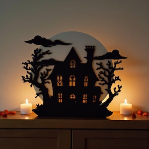 Pu&ograve; includere: Silhouette nera di una casa infestata con sfondo di luna piena, alberi e nuvole. Due candele accese affiancano la casa su una superficie di legno, creando una scena di Halloween spettrale.