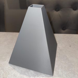 May include: A gray, pyramid-shaped lampshade with a square opening at the top. The lampshade has a smooth, matte finish and is made of a solid material. The design is modern and geometric.