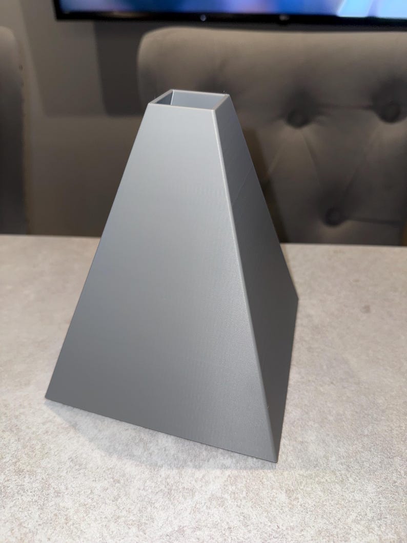 May include: A gray, pyramid-shaped lampshade with a square opening at the top. The lampshade has a smooth, matte finish and is made of a solid material. The design is modern and geometric.
