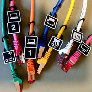 May include: A collection of colorful Ethernet cables, each with a different color and attached black cable ties. Each cable has a white label with a black icon, such as a computer, printer, or Wi-Fi symbol. Some labels have numbers.