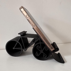 May include: A black bicycle-shaped phone stand holds a smartphone. The stand has two large circular wheels and a frame that resembles a bicycle. The phone is in a clear case with a gold-colored frame.