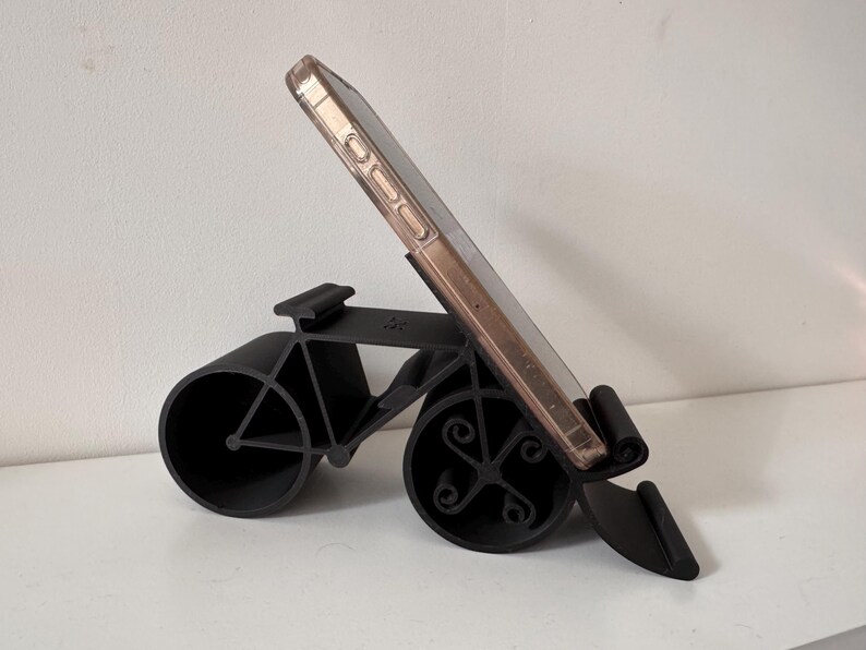 May include: A black bicycle-shaped phone stand holds a smartphone. The stand has two large circular wheels and a frame that resembles a bicycle. The phone is in a clear case with a gold-colored frame.