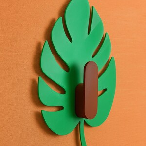 以下が含まれることがあります： オレンジ色のテクスチャード加工された壁に取り付けられた、茶色のフックが付いた緑色の葉の形をしたウォールフック。葉には切り抜きのある詳細なデザインがあり、フックは対照的な色です。