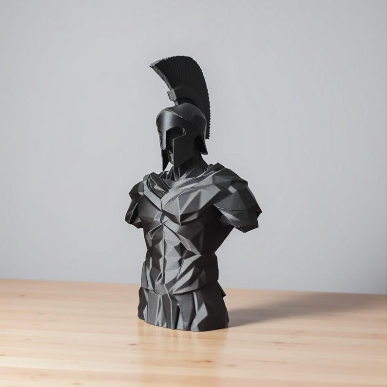 May include: A black sculpture of a Spartan warrior's bust, with a helmet and crest, and geometric armour. The artwork is displayed on a wooden surface. The sculpture has a geometric, faceted design.