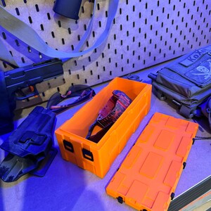 May include: Bright orange rectangular storage box with a matching lid, open to reveal a pair of camouflage-patterned glasses. A black holster and a handgun are visible in the background. The box is designed with a series of rectangular indentations.