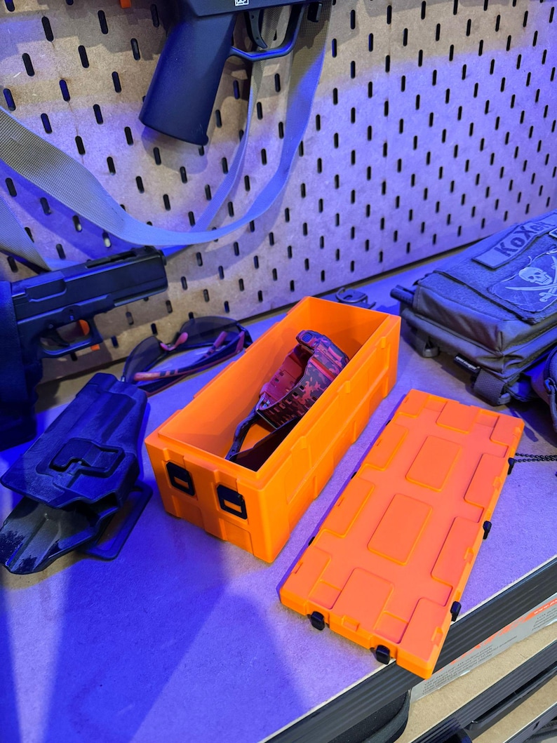 May include: Bright orange rectangular storage box with a matching lid, open to reveal a pair of camouflage-patterned glasses. A black holster and a handgun are visible in the background. The box is designed with a series of rectangular indentations.