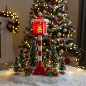 Op de afbeelding: Een feestelijke kerstsc&egrave;ne met een rood-witte vuurtorenlantaarn die een warme gloed uitstraalt. Miniatuur kerstbomen en ingepakte cadeaus omringen de lantaarn, geplaatst op een besneeuwd oppervlak. Een versierde kerstboom en open haard zijn op de achtergrond te zien.