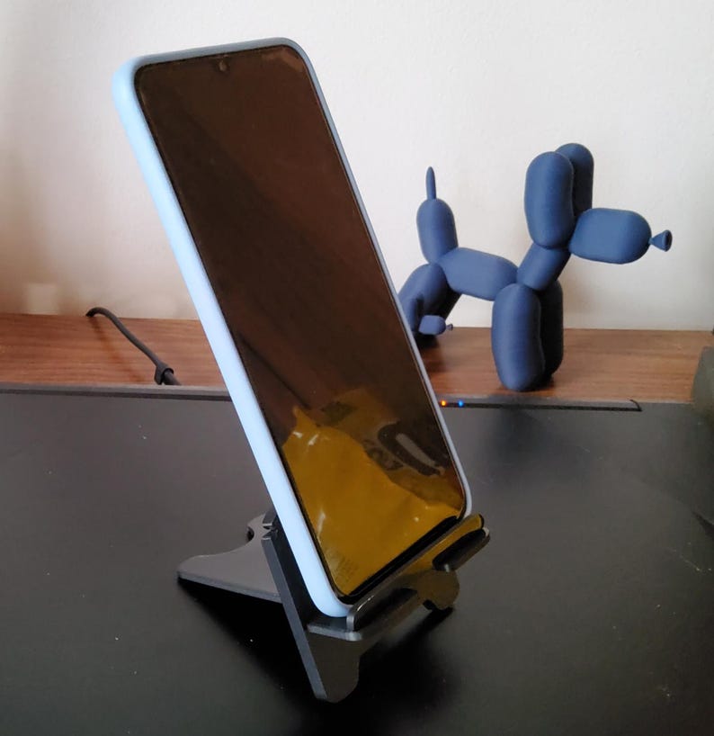 May include: A black phone stand holding a smartphone with a light blue case. A blue balloon dog sculpture is in the background. The phone stand is on a black surface.
