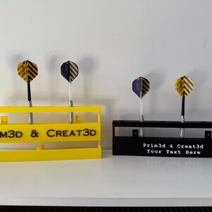 Puede incluir: Dos soportes para dardos, uno amarillo y otro negro, cada uno con tres dardos. El soporte amarillo tiene el texto "PRIM3D & CREAT3D". El soporte negro tiene el texto "Prim3d & Creat3d Your Text Here". Los dardos tienen plumas a rayas amarillas y negras o azules y negras.