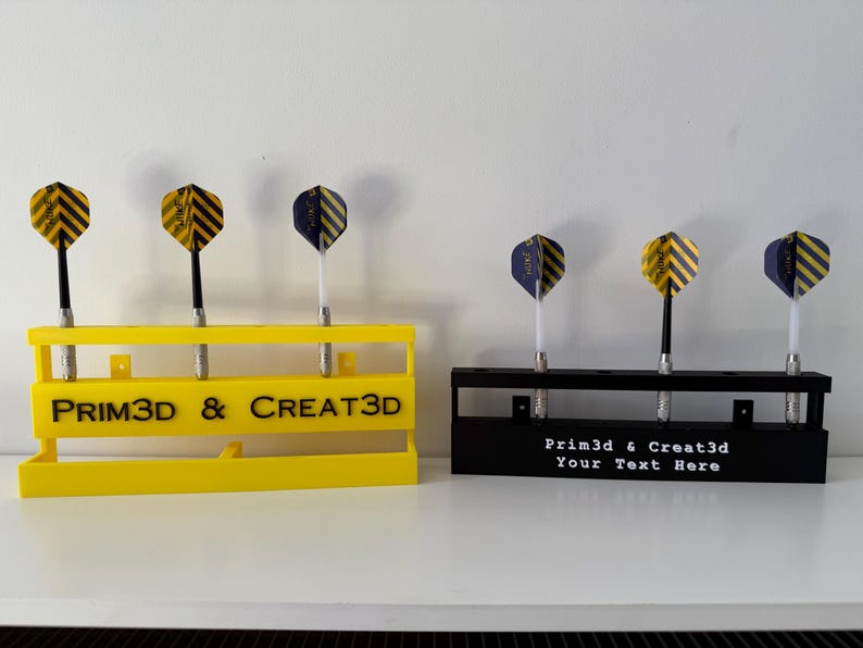 Puede incluir: Dos soportes para dardos, uno amarillo y otro negro, cada uno con tres dardos. El soporte amarillo tiene el texto "PRIM3D & CREAT3D". El soporte negro tiene el texto "Prim3d & Creat3d Your Text Here". Los dardos tienen plumas a rayas amarillas y negras o azules y negras.