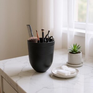 May include: A black, cylindrical container with horizontal grooves holds a comb, makeup brushes, and scissors. A small succulent in a gray pot sits nearby, along with a small, white plate with two white bars. The items are arranged on a white marble surface.