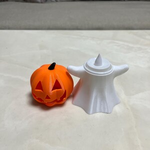 May include: An orange pumpkin with a carved face and a black stem sits next to a white ghost figurine. The Halloween-themed decorations are made of plastic and are suitable for indoor use.