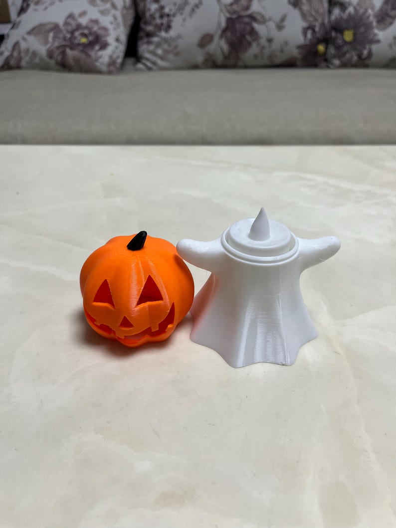 May include: An orange pumpkin with a carved face and a black stem sits next to a white ghost figurine. The Halloween-themed decorations are made of plastic and are suitable for indoor use.