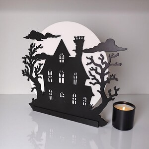 Pu&ograve; includere: Silhouette nera di una casa infestata con una luna bianca, alberi e nuvole. A destra c'&egrave; una candela nera con uno stoppino acceso. La casa ha diverse finestre e un camino, creando una spettrale decorazione di Halloween.