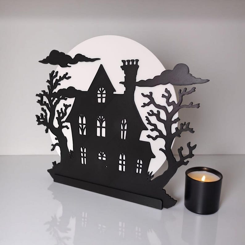 Pu&ograve; includere: Silhouette nera di una casa infestata con una luna bianca, alberi e nuvole. A destra c'&egrave; una candela nera con uno stoppino acceso. La casa ha diverse finestre e un camino, creando una spettrale decorazione di Halloween.