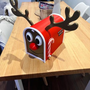 May include: A red mailbox with reindeer antlers, a red nose, and large cartoon eyes. A candy cane is attached to the side. The mailbox is on a wooden table, perfect for Christmas decorations.