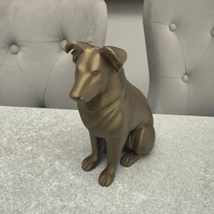 May include: A gold-colored dog figurine in a seated position. The sculpture has detailed features, including the dog's ears, paws, and facial expression. The dog is sitting on a light-colored surface.