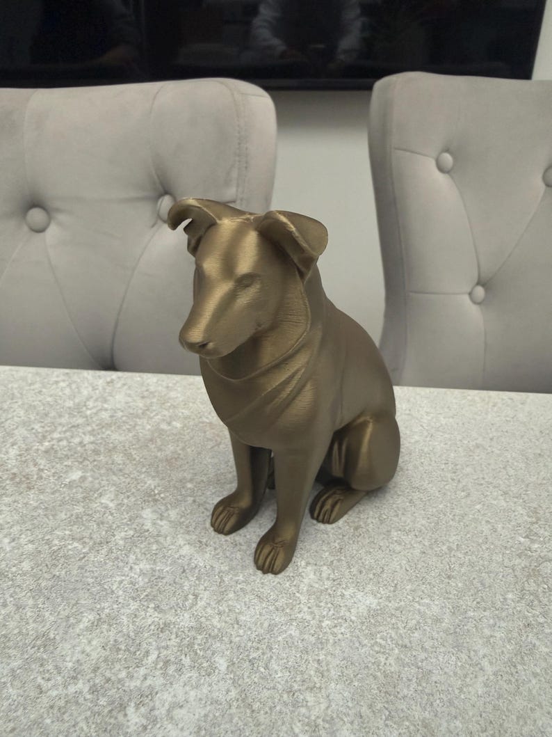 May include: A gold-colored dog figurine in a seated position. The sculpture has detailed features, including the dog's ears, paws, and facial expression. The dog is sitting on a light-colored surface.