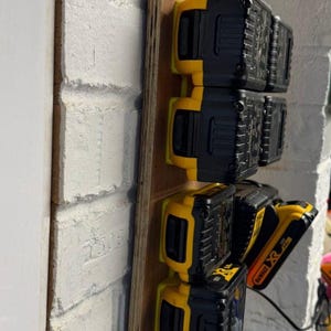 May include: A wooden mounting board holds several black and yellow power tool batteries. The batteries are arranged vertically, with the DeWalt logo visible on one. The board is mounted on a white brick wall.
