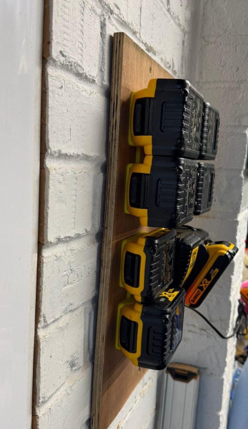 May include: A wooden mounting board holds several black and yellow power tool batteries. The batteries are arranged vertically, with the DeWalt logo visible on one. The board is mounted on a white brick wall.