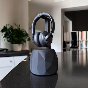 May include: A black over-ear headphone set rests on a matching black headphone stand. The stand has a geometric, faceted design. The headphones are suspended above the stand, showcasing their sleek, modern aesthetic. The background is a blurred interior.