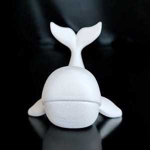 May include: A white ceramic whale figurine with a textured surface. The whale is depicted with its tail raised and flippers extended. The figurine is set against a black background, creating a contrast that highlights its form.