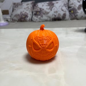 May include: An orange pumpkin with a carved Spider-Man face design. The pumpkin features a detailed web pattern and eye shapes, perfect for Halloween decorations. The pumpkin is sitting on a white surface.