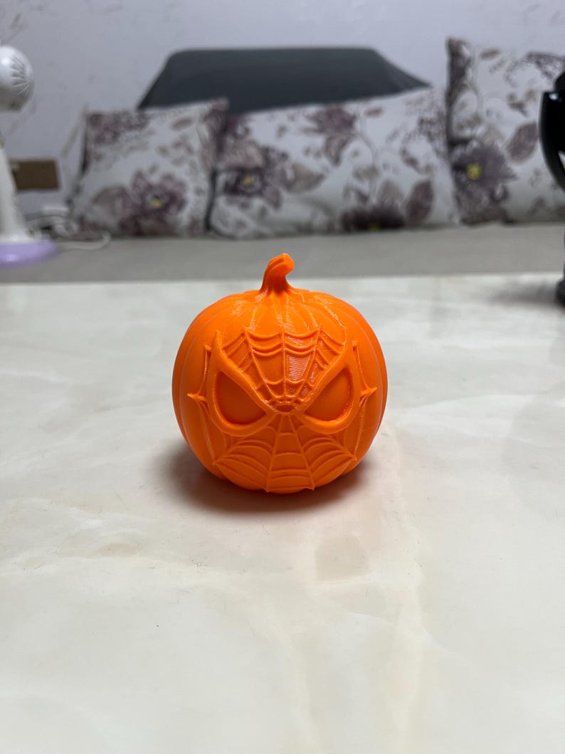 May include: An orange pumpkin with a carved Spider-Man face design. The pumpkin features a detailed web pattern and eye shapes, perfect for Halloween decorations. The pumpkin is sitting on a white surface.