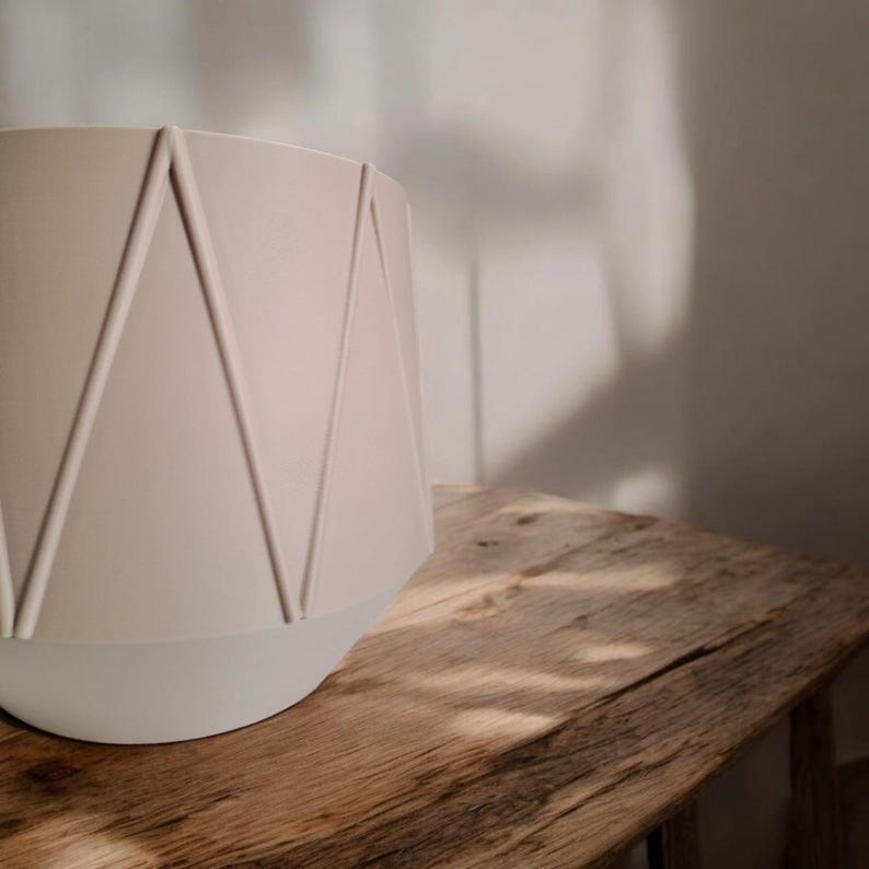 May include: A beige geometric planter with a white base sits on a rustic wooden table. The planter has a series of raised, angular lines that create a geometric pattern. The neutral colors and simple design suggest a modern aesthetic.