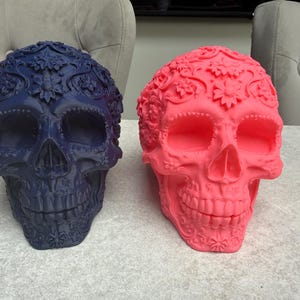 May include: Two decorative skull sculptures. One is navy blue, the other is bright pink. Both skulls have floral and ornamental details. The skulls are placed side by side on a light gray surface.