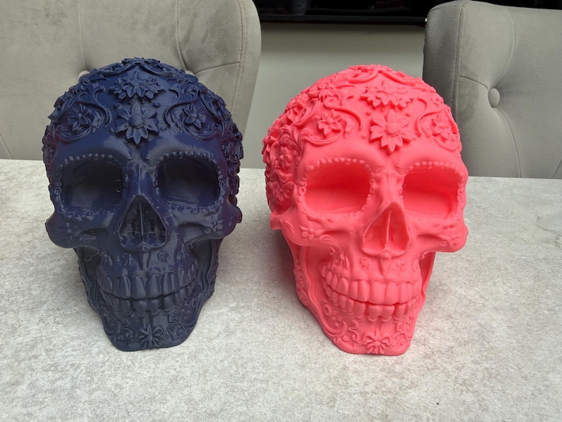 May include: Two decorative skull sculptures. One is navy blue, the other is bright pink. Both skulls have floral and ornamental details. The skulls are placed side by side on a light gray surface.